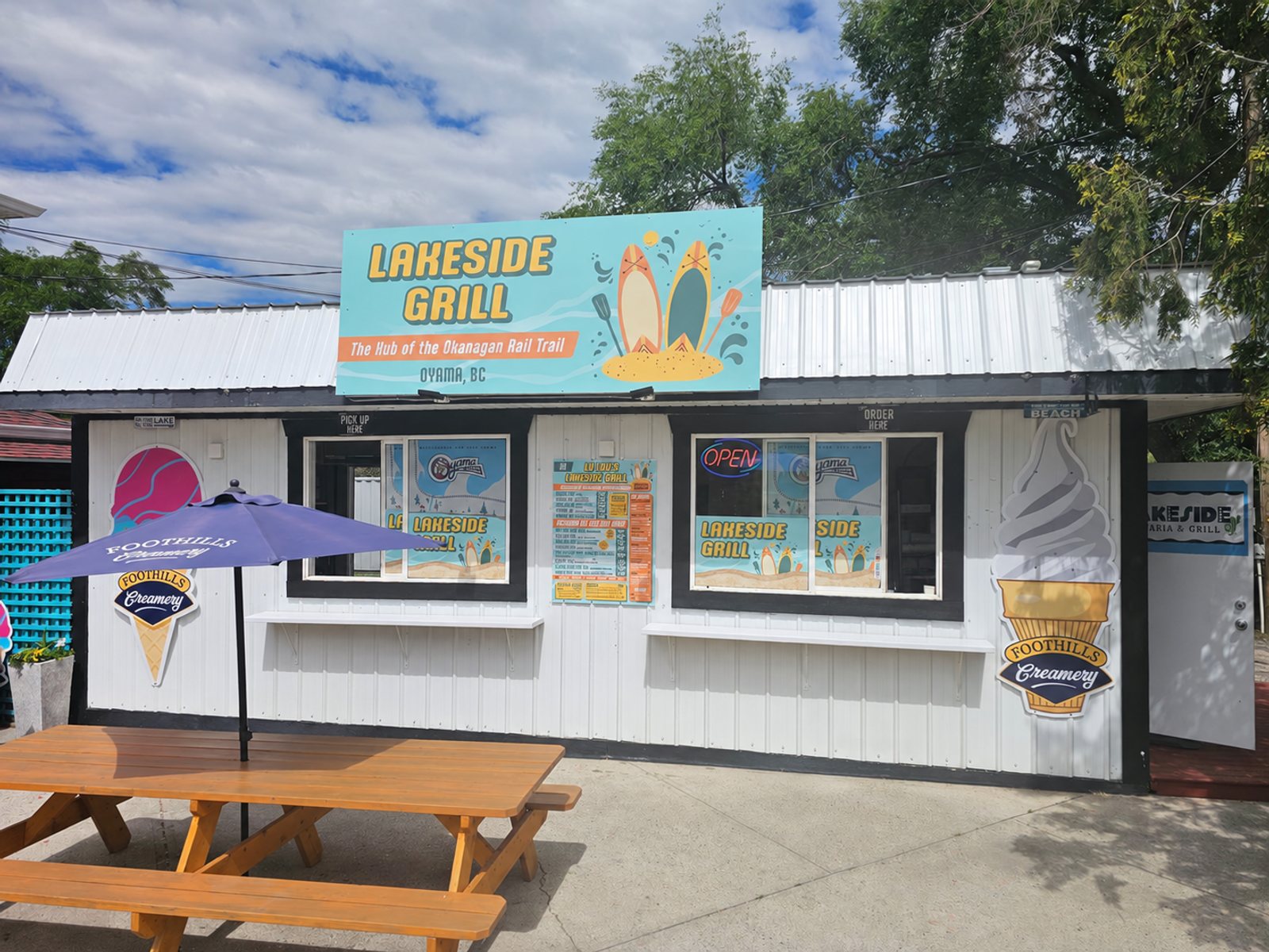 The Lu Lou's grill stand on the beach in Oyama, BC, with the orange and teal sign visible above the wooden deck